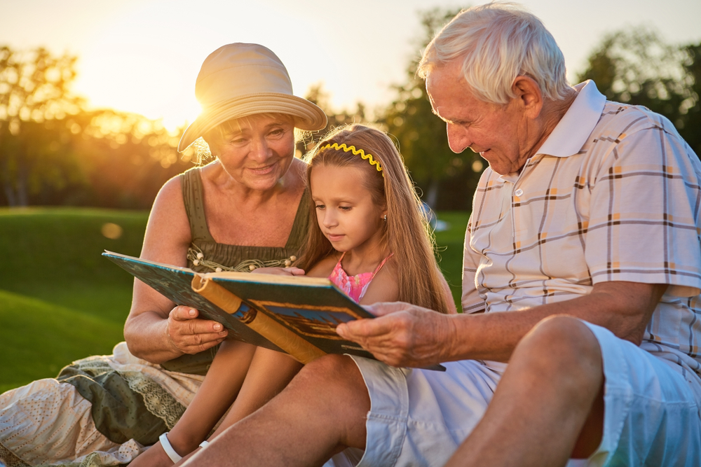 Elderly couple reading a book with their granddaughter outdoors in a sunlit park.