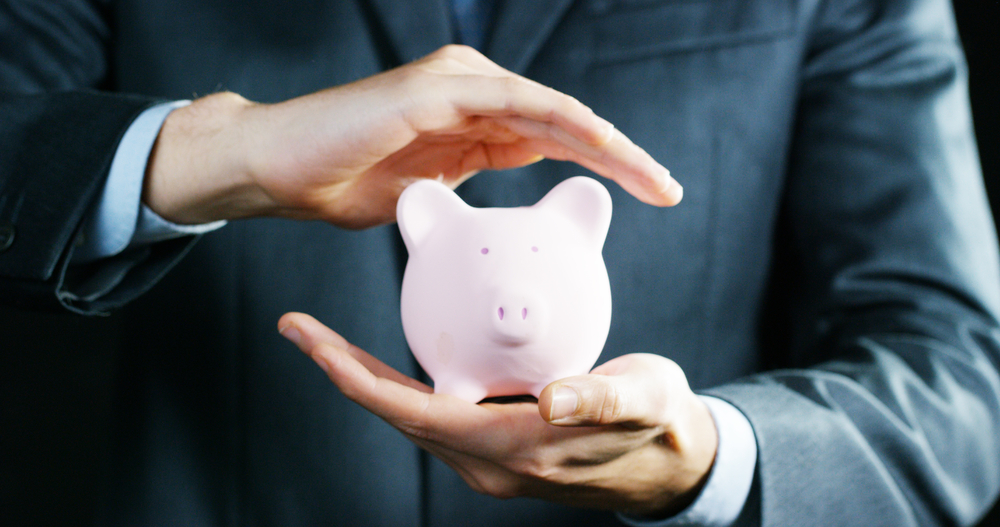 Person in suit holding and protecting a small pink piggy bank with hands.