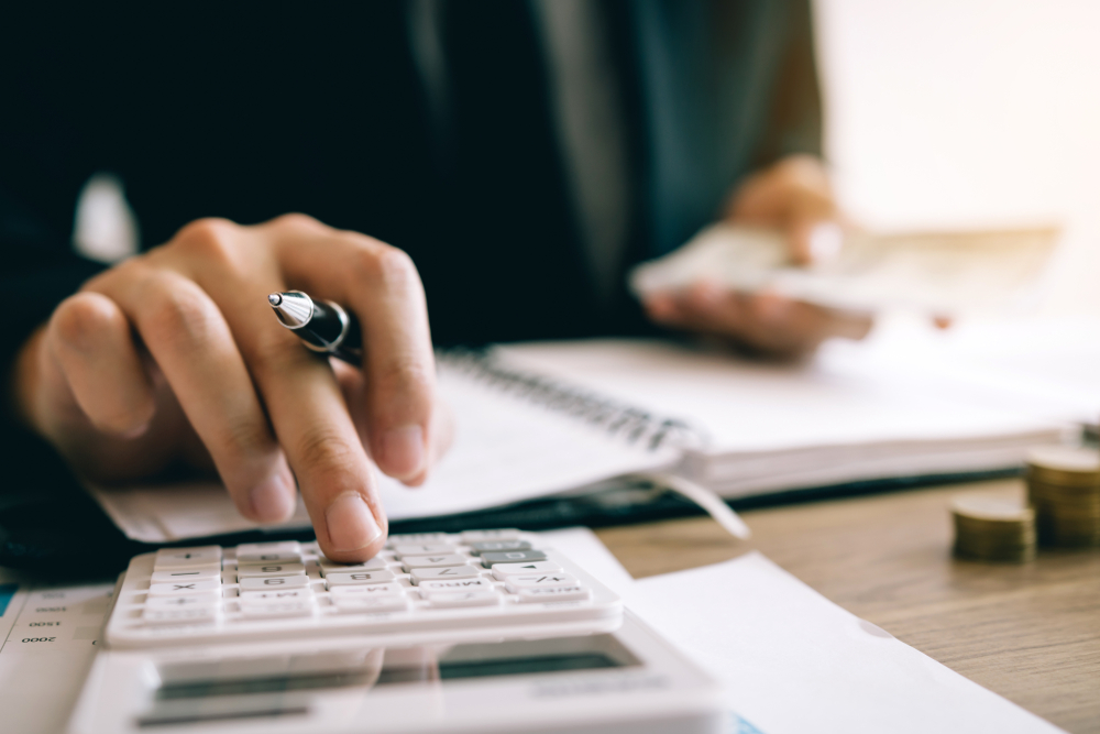 Person using a calculator with one hand while holding a pen and counting money, with an open notebook on a desk.