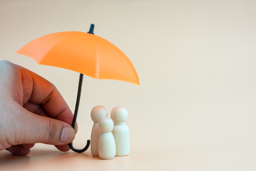 Hand holding an orange miniature umbrella over three wooden peg figures on a light background.
