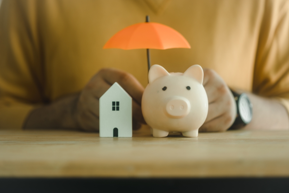 A person in a mustard sweater holds a small orange umbrella over a white piggy bank and a miniature white house on a wooden table.