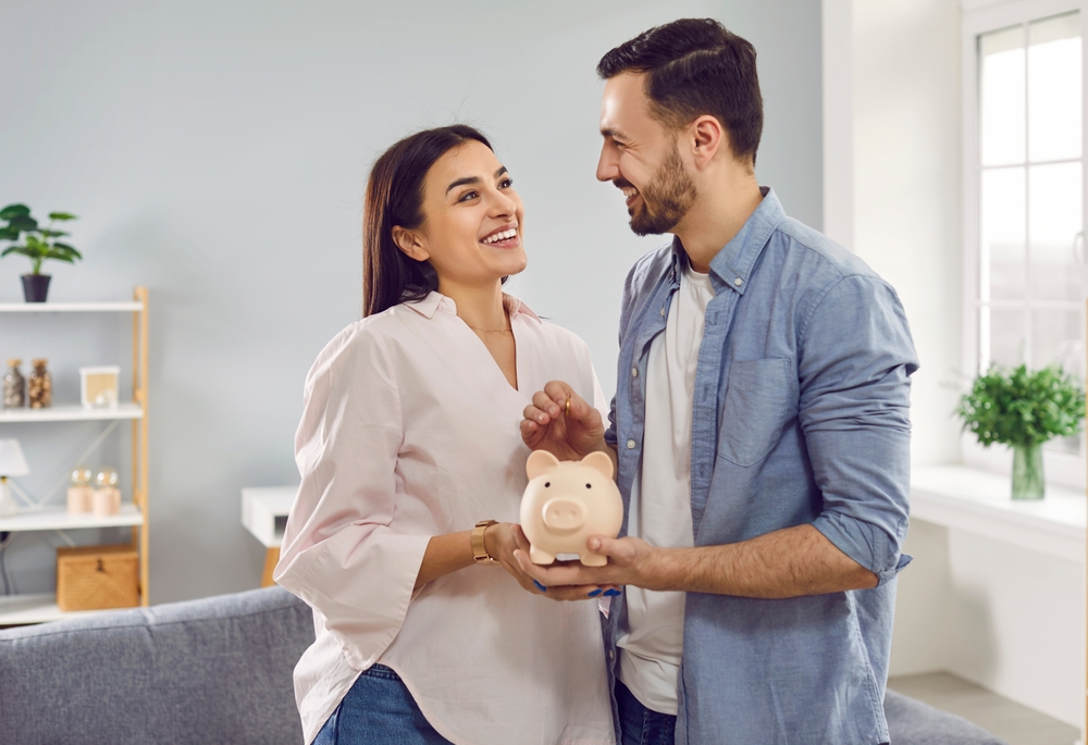 Happy couple holding a piggy bank, smiling at each other in a bright living room.