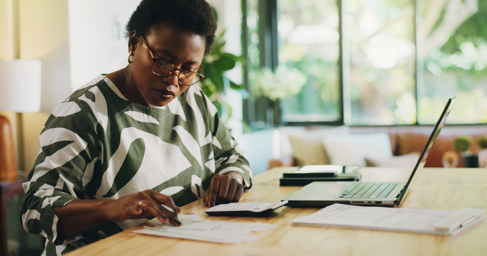 Woman wearing glasses and patterned shirt working on paperwork with a calculator and laptop at a wooden table in a bright room.