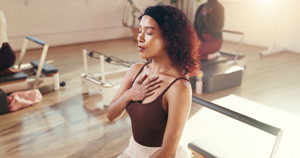 Woman with curly hair and headband sitting with eyes closed, placing hand on chest during a Pilates class.