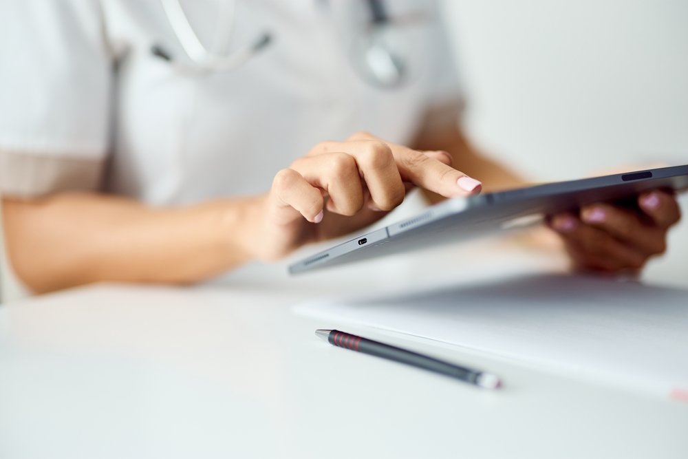 Medical professional using a tablet with a stethoscope around their neck and a pen on the desk.