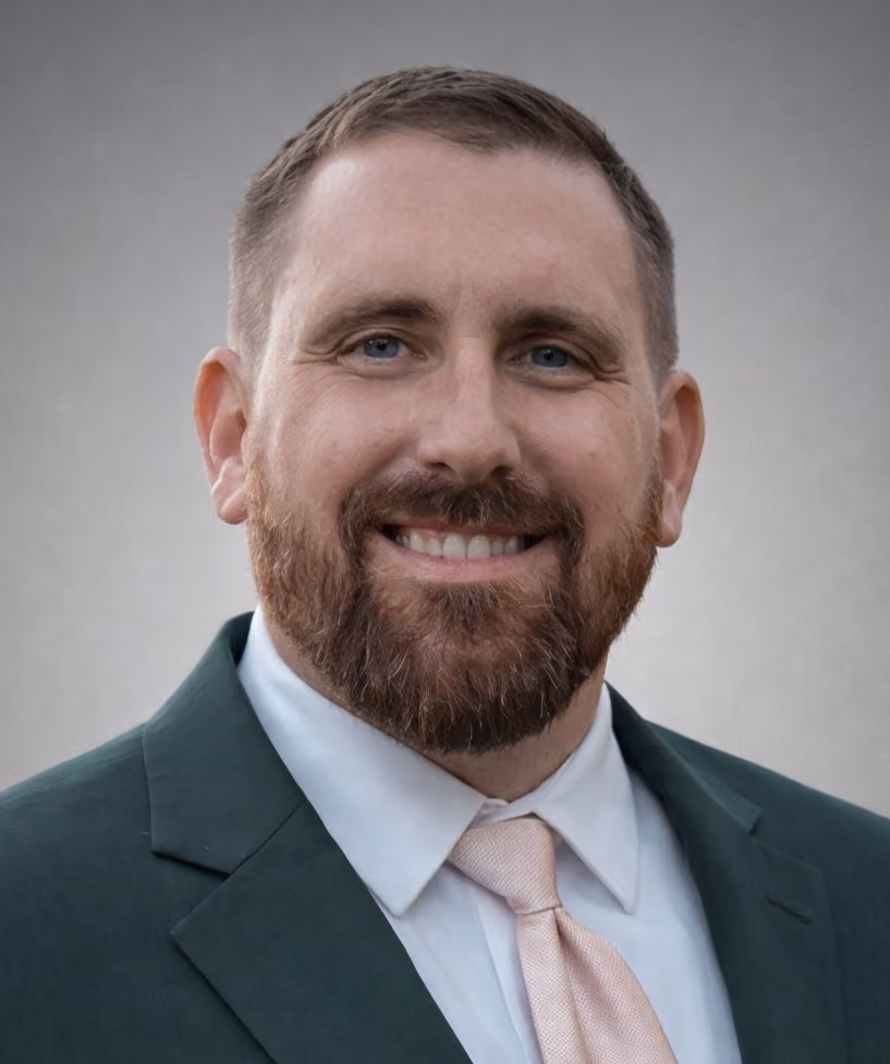 Smiling man with short brown hair and beard, wearing a dark green suit jacket, white shirt, and light pink tie, against a plain gray background.