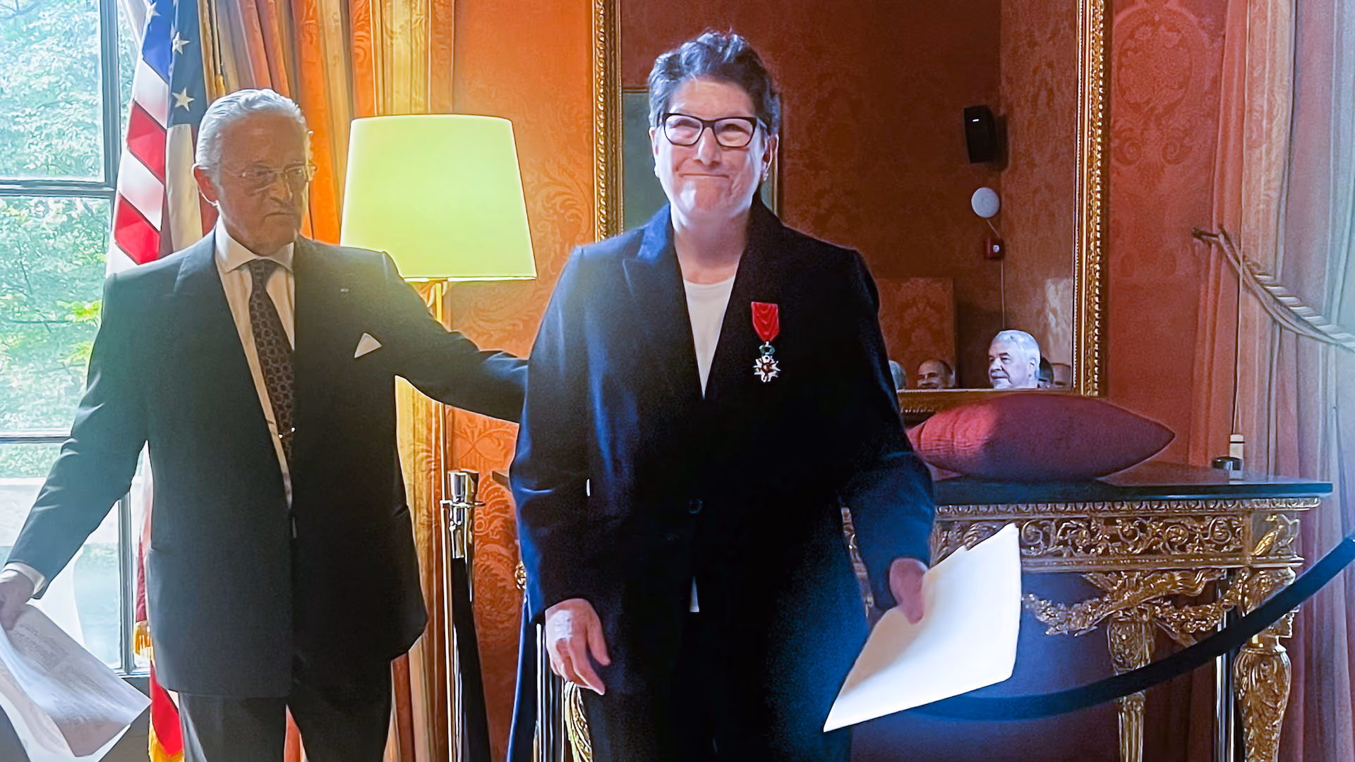 A middle-aged woman in a suit stands in an ornate room smiling as she faces the camera. Pinned on her lapel is the medal of the National Order of the Legion of Honor. A man in a suit stands behind her.