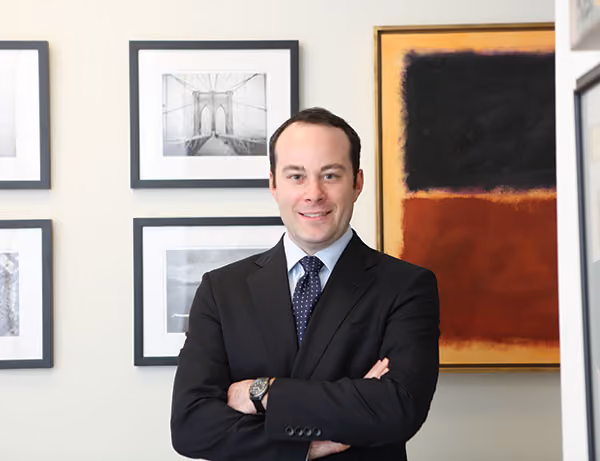 Luke Nikas, wearing a suit and crossing his arms, stands in front of a wall with framed photography and artwork.