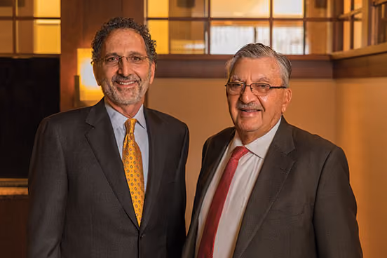 Norman and Morris Massry stand next to each other and smile in a room with a warm orange glow.