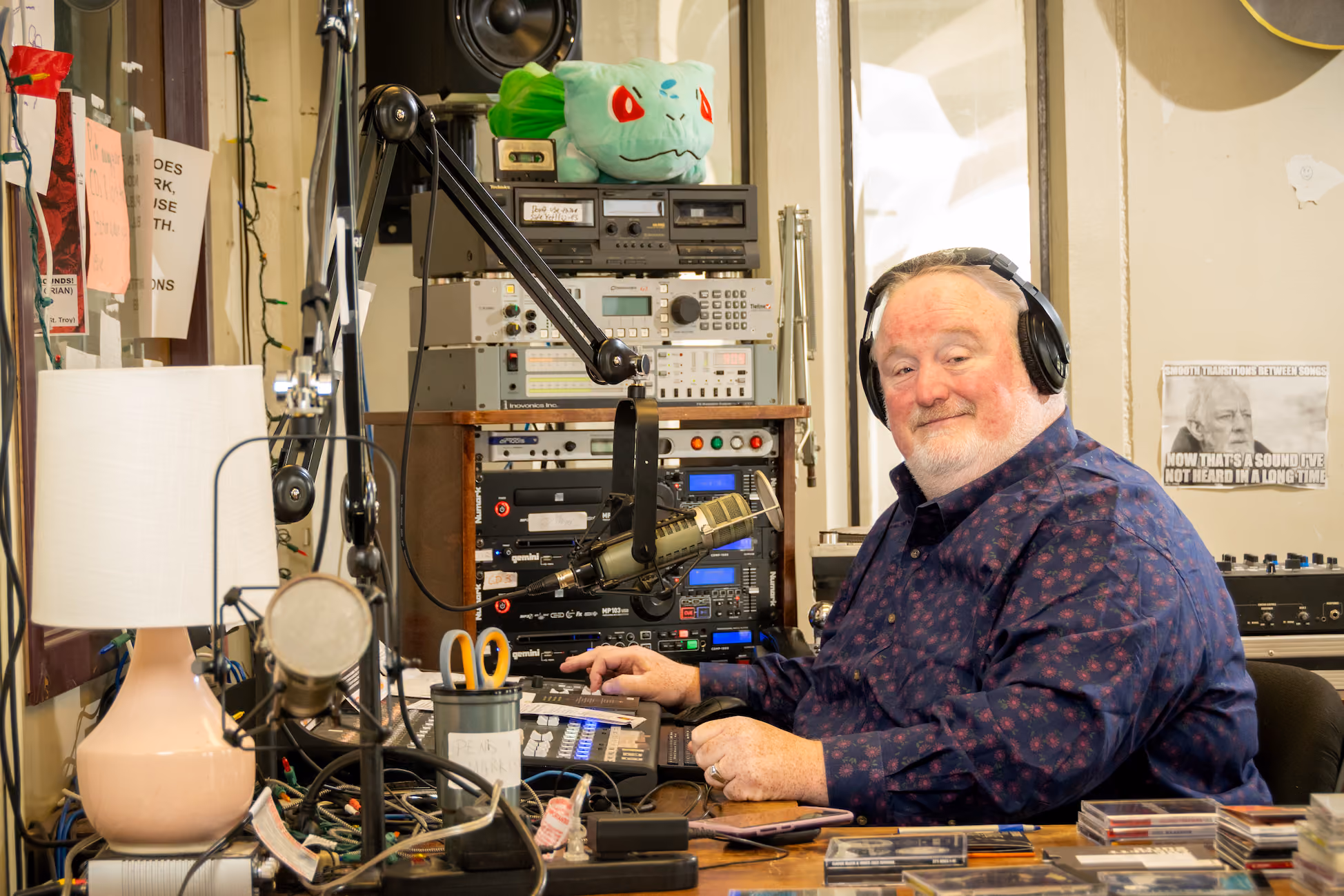 Bill McCann, a white middle-aged male, smiles while wearing headphones and posing behind the audio board of WCDB 90.9FM radio.