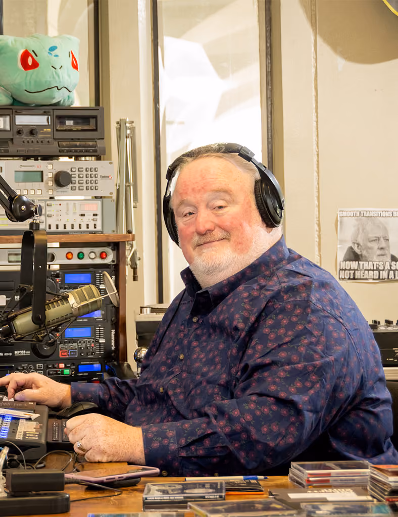 Bill McCann, a white middle-aged male, smiles while wearing headphones and posing behind the audio board of WCDB 90.9FM radio.