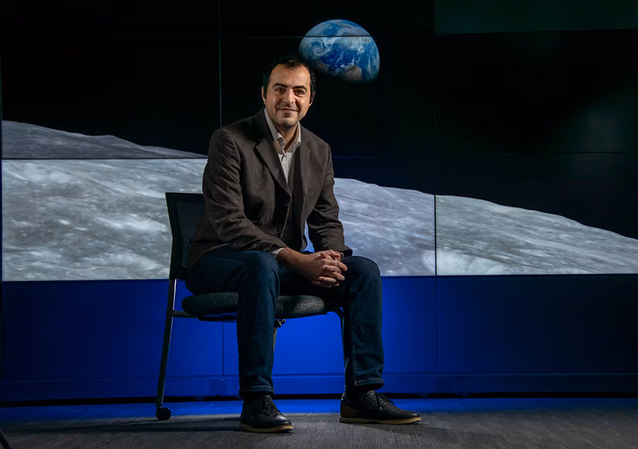 UAlbany professor Mustafa Aksoy sits in front of a multi-screen display with a NASA image of the Earth rising over the horizon of the moon.