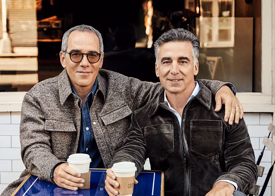The Lipman brothers sit at a small table outdoors with cups of coffee.