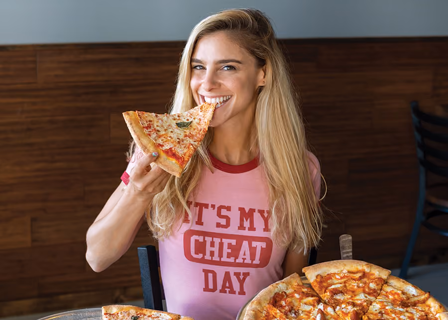 Jessica wearing a pink t-shirt with the text "It's my cheat day" and eating a large slice of pizza.