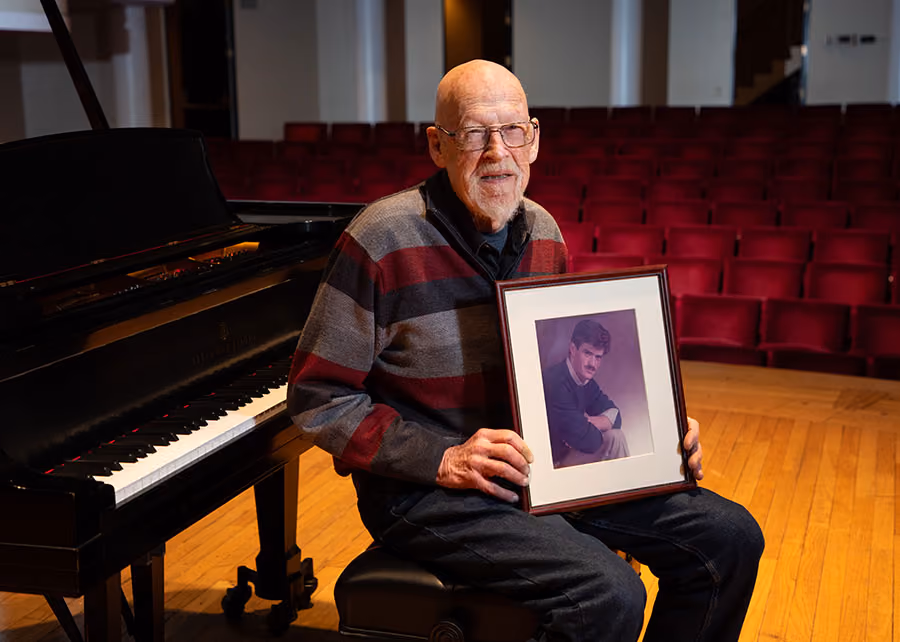 George Hastings sitting on a piano bench in an auditorium and holding a framed photo of his son Douglas.