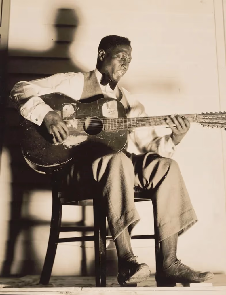 An archival black and white image of an Africa-American musician, seated on a chair, playing an acoustic guitar.