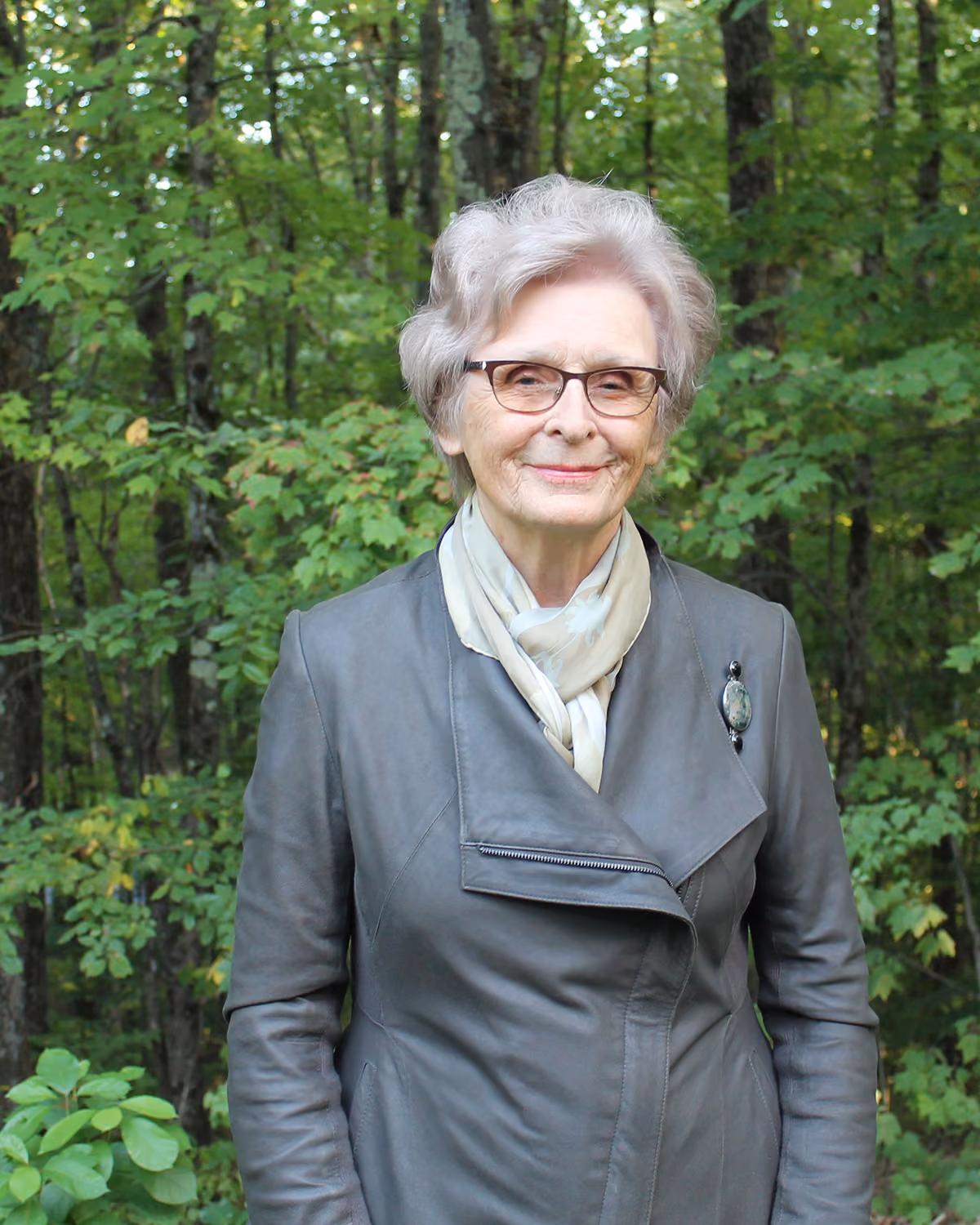 Ellen Jabur, a white female senior citizen, dressed in a gray jacket and cream neck scarf, poses in front of a wooded background
