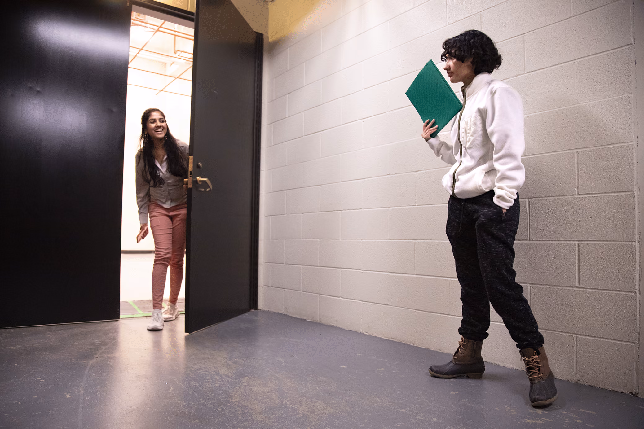 A female student opens a door to greet another student holding a binder.