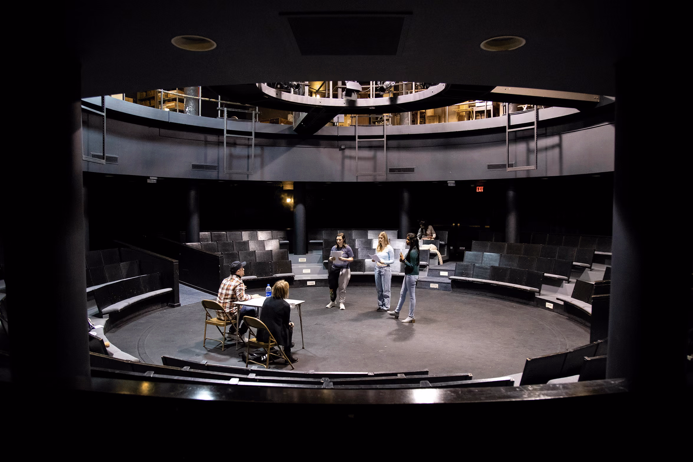 In a circular theatre space, student actors stand and address two people sitting behing a folding table.