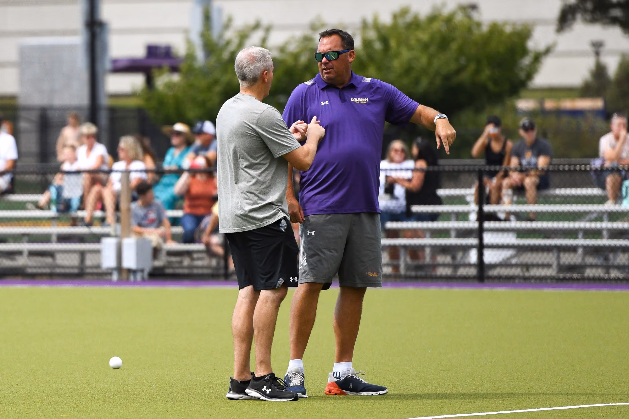 Two white male coaches are in a discussion on a field hockey field.