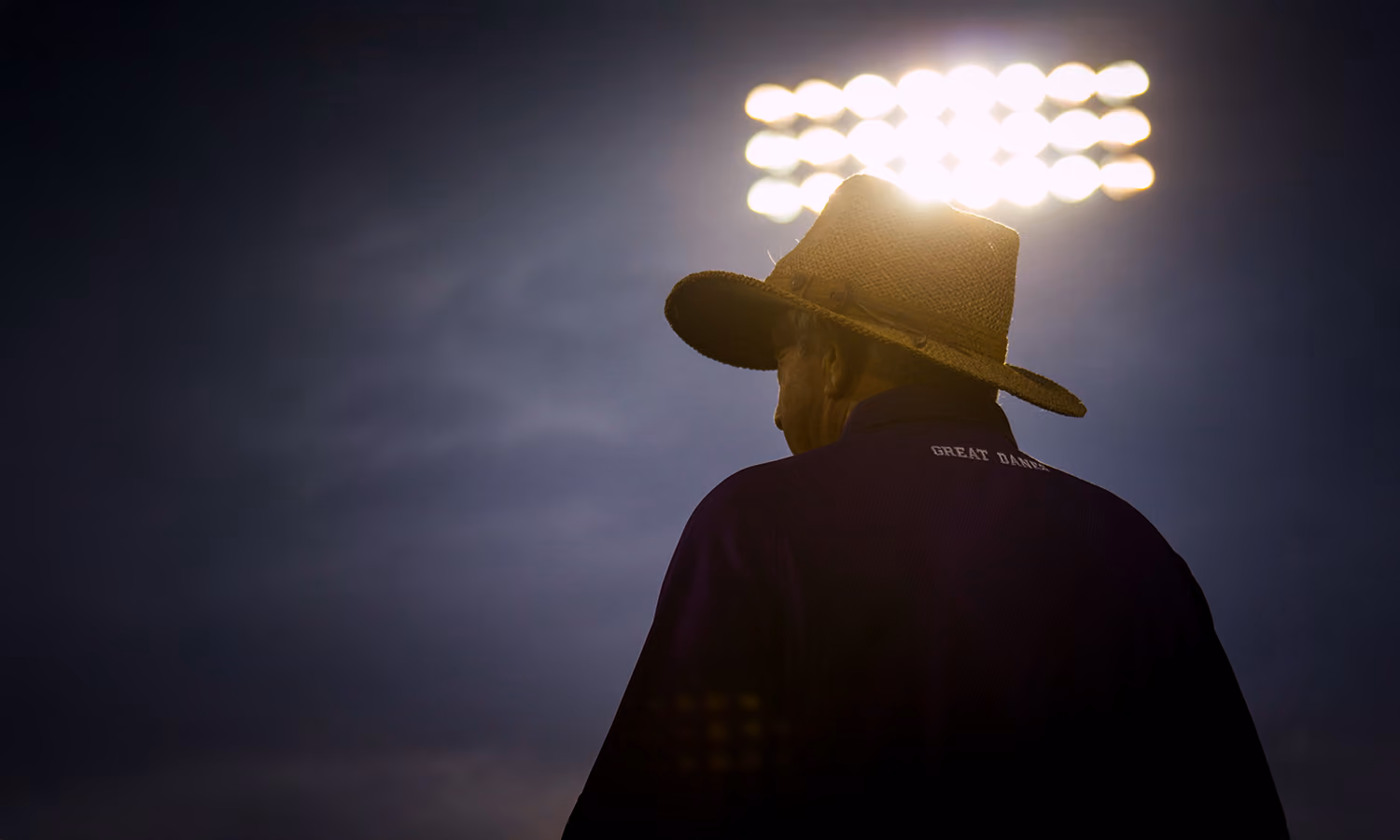 silhouette of a man wearing a Stetson cowboy hat facing away from camera as a large stadium light glows in the background