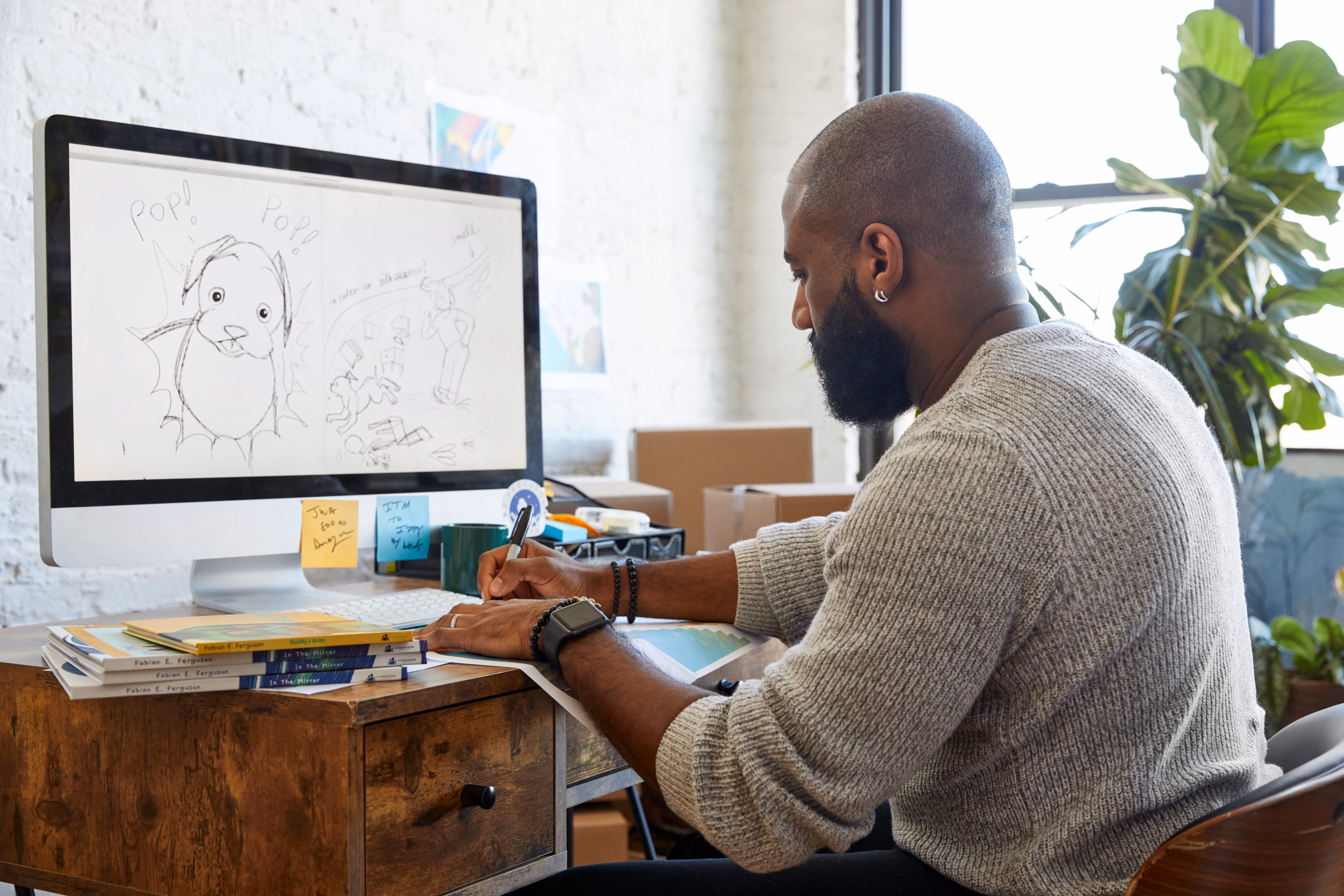 An African American man sits in front of a computer sketching with marker while sketched illustrations appear on a computer screen in front o fhim.