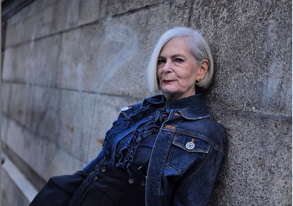 Lyn Slater wearing a black jacket leans against a brick wall.