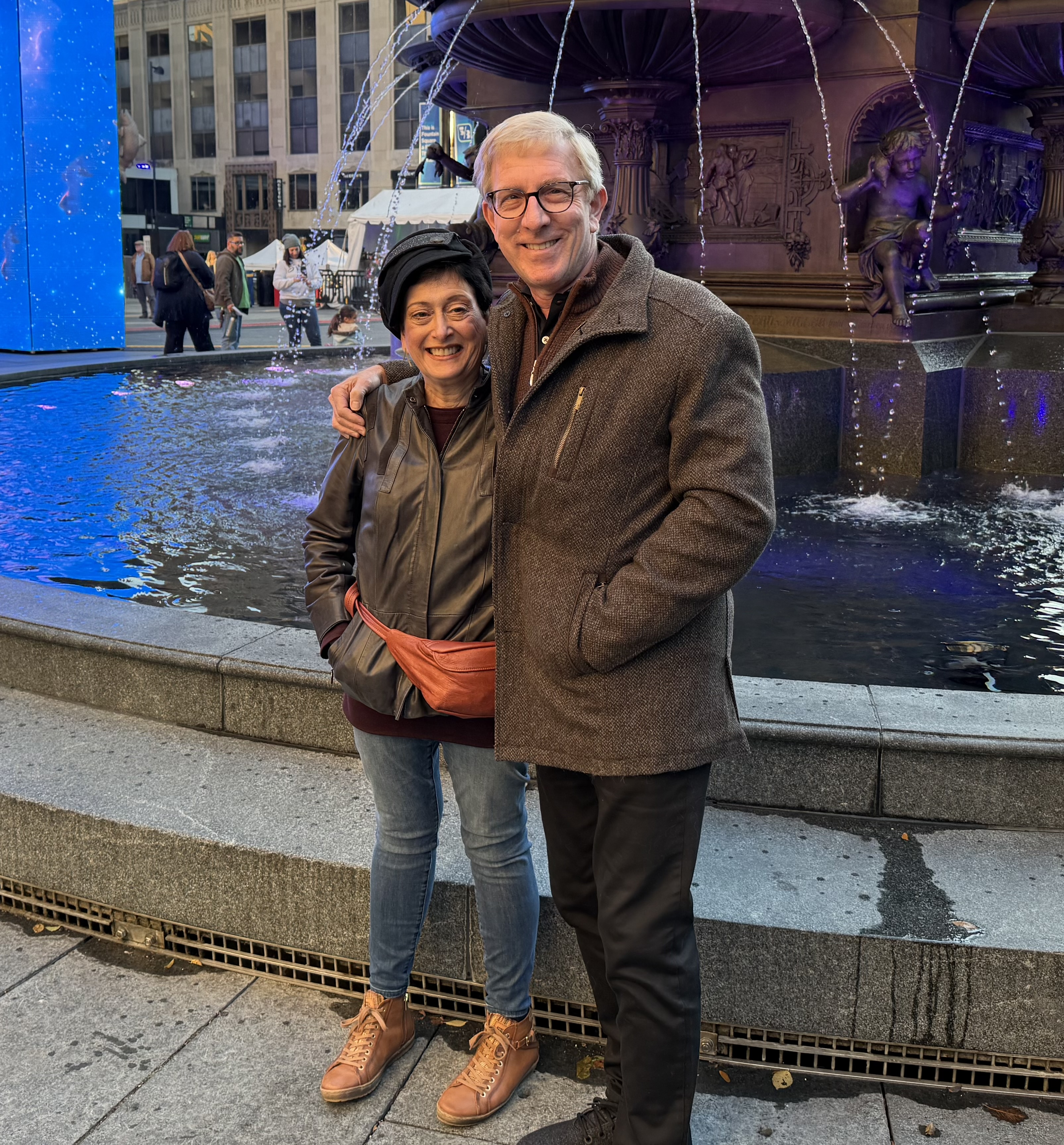 Jan and Mark Rosenbaum, a white couple of about middle age, stand smiling in front large fountain in a city.