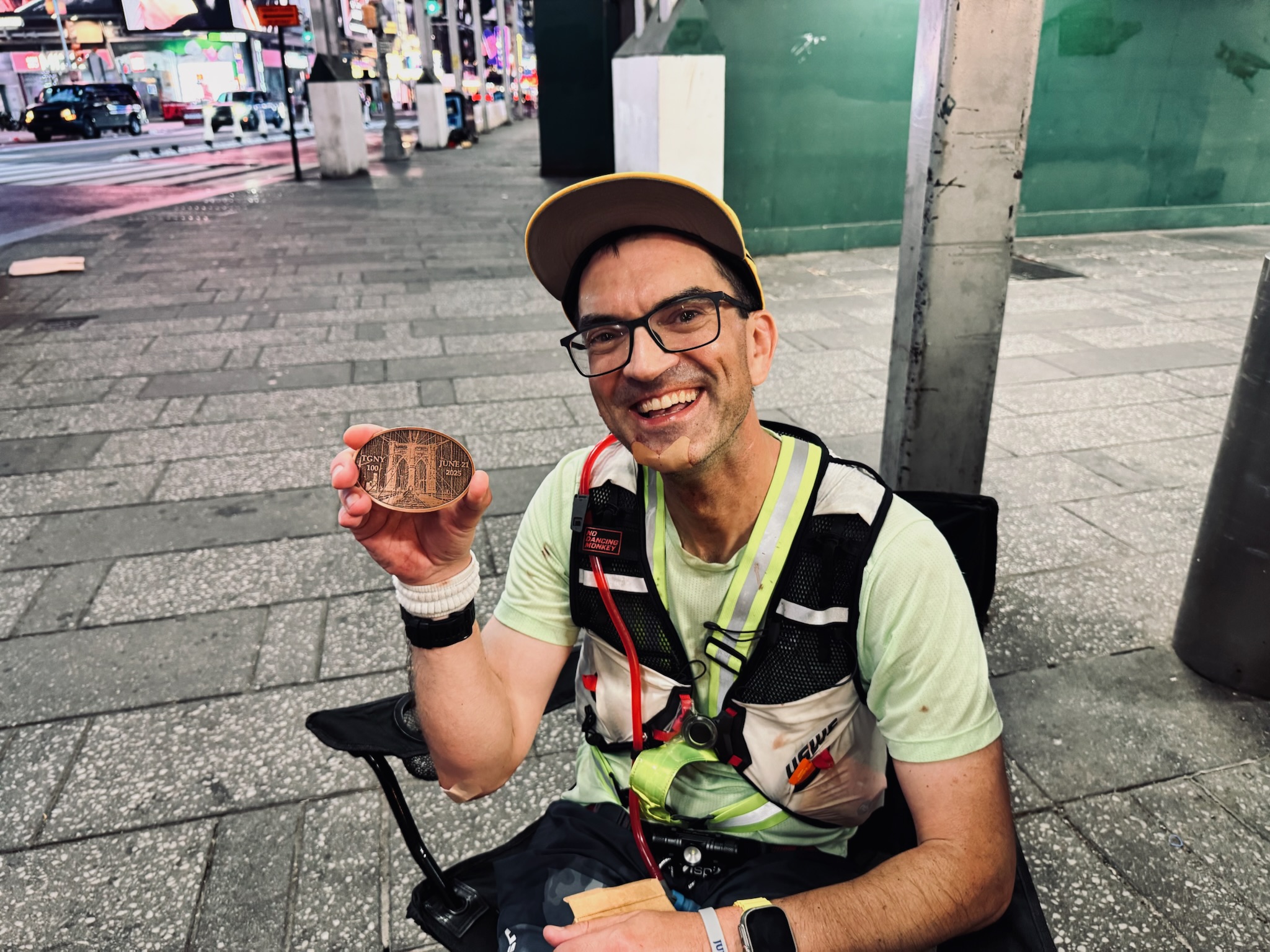 A male runner smiles broadly and holds up a bronze medallion while sitting in a folding chair.