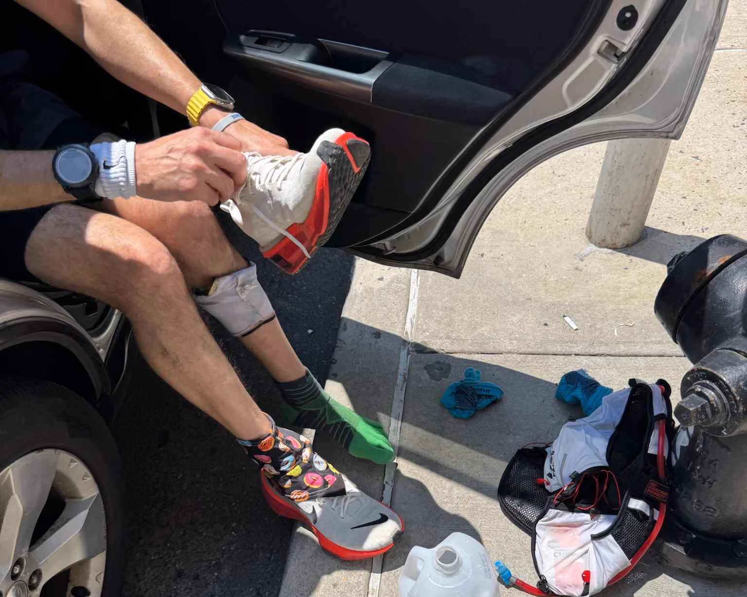 shot of a man's arms putting on a running show while sitting halfway into a car. An open gallon of water and clothing are on the ground.