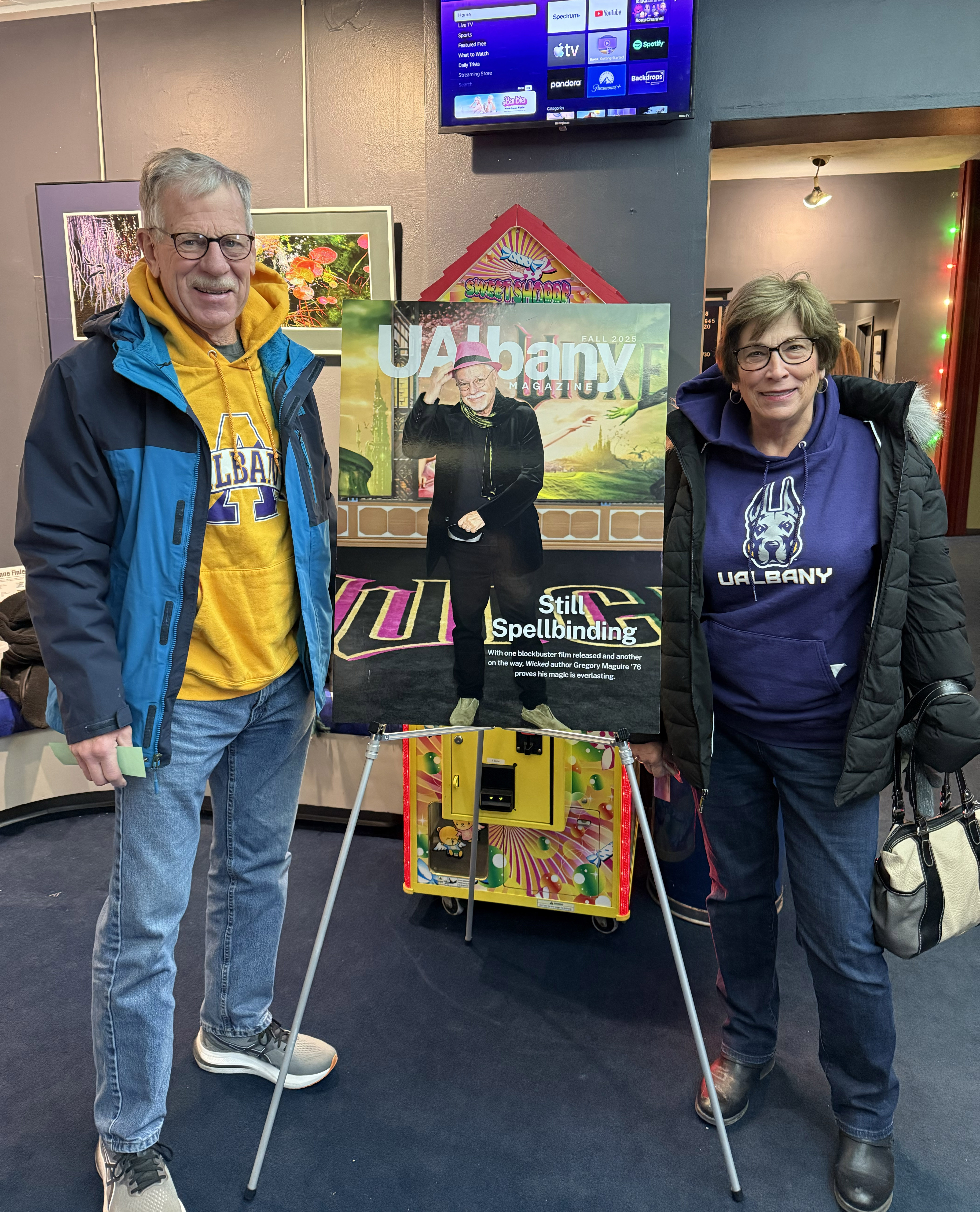 Two people stand beside a poster of the UAlbany Magazine cover featuring Gregory Maguire.