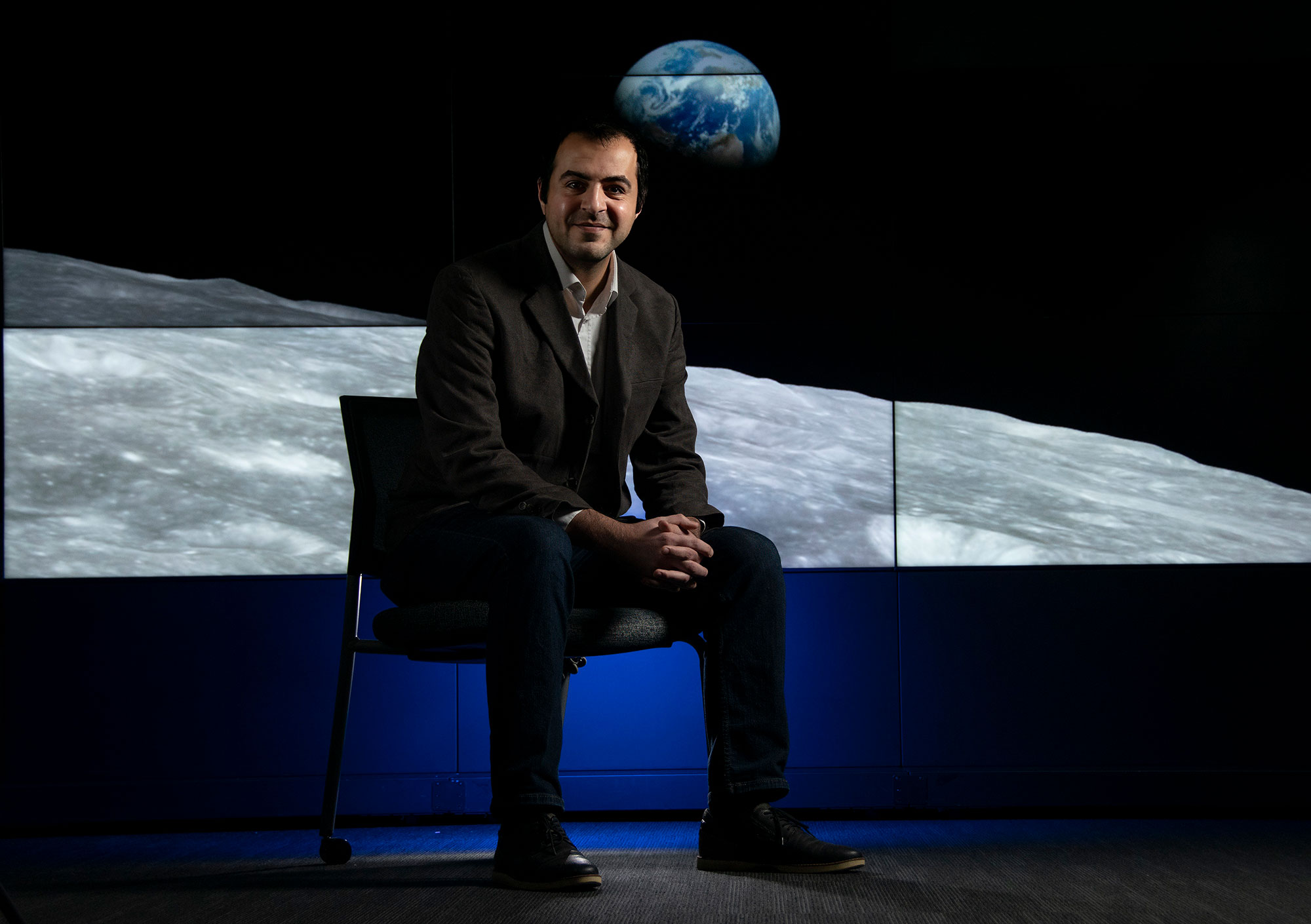 UAlbany professor Mustafa Aksoy sits in front of a multi-screen display with a NASA image of the Earth rising over the horizon of the moon.