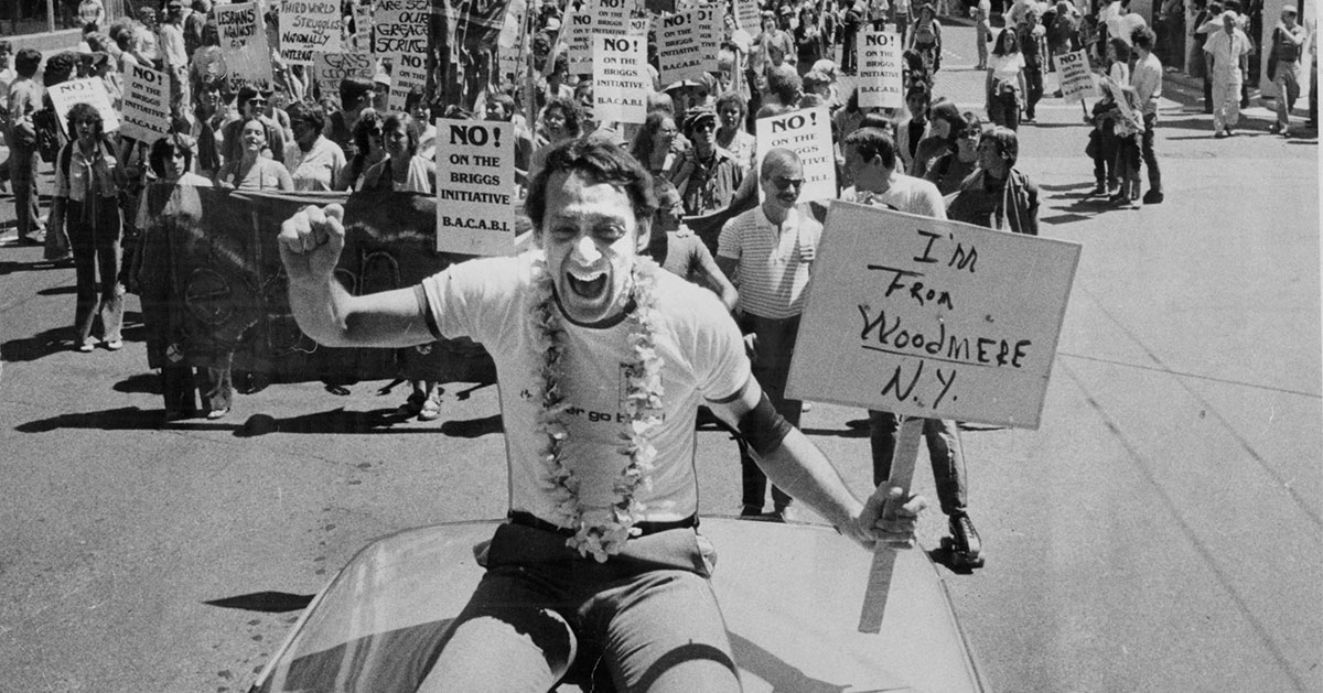 Harvey Milk sits on top of a convertible and cheers while leading a parade and holding a sign that reads "I'm From Woodmere N.Y."