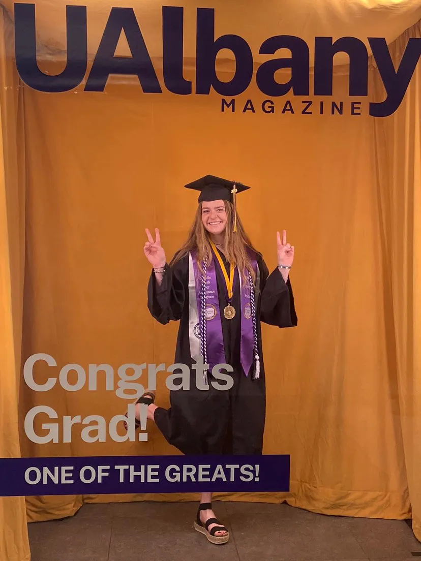 A graduate holds up hands in two peace signs while posing for a photo in the UAlbany Magazine photo booth.