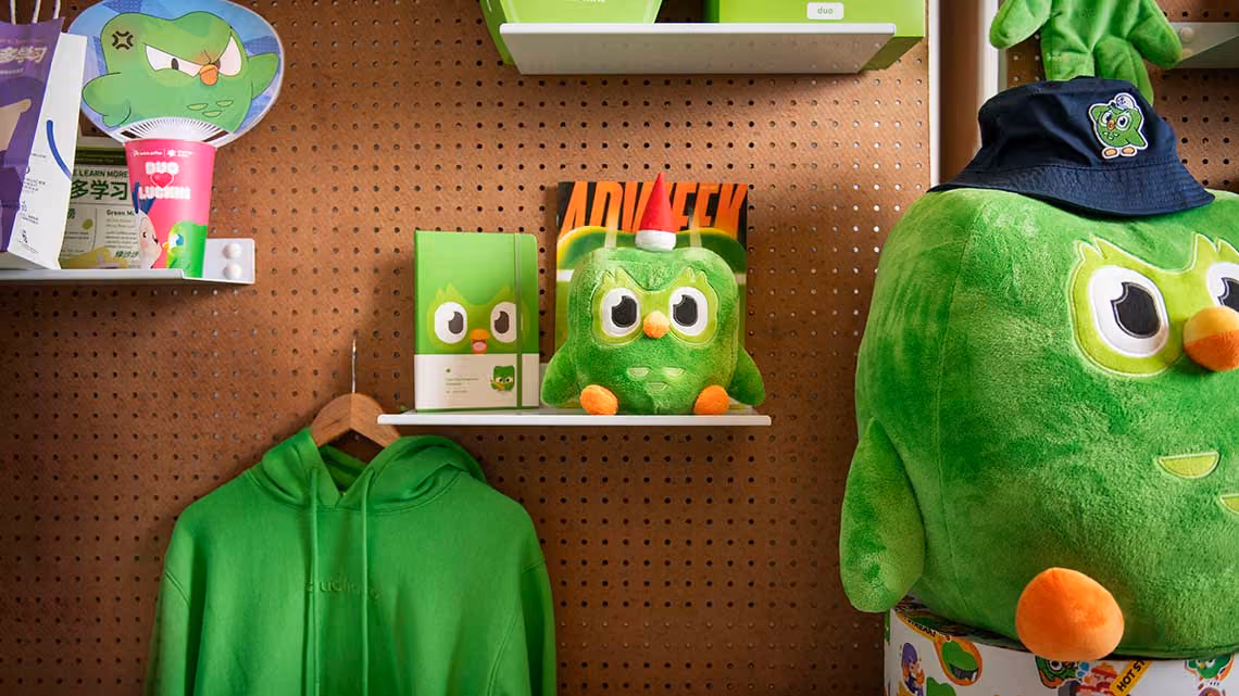 A wall of Duolingo merchandise including Duo plushies and a green hoodie.
