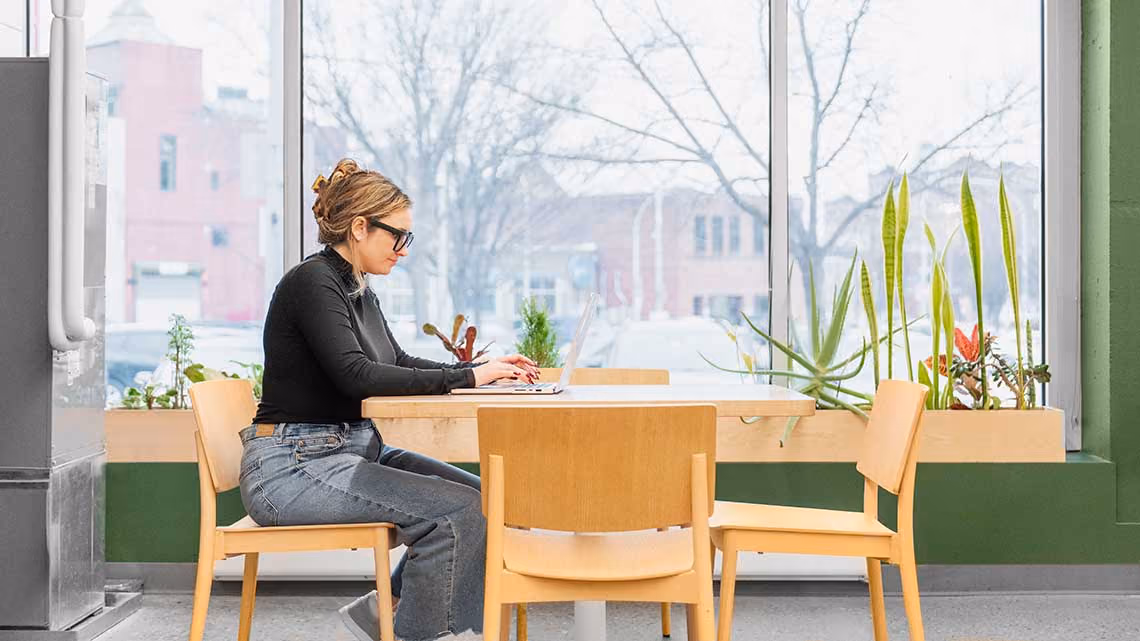 Adele sitting alone at a light wooden table in front of a large window.