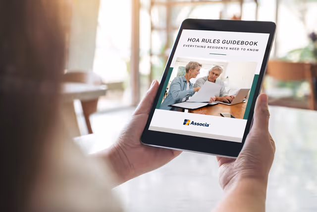 A person holds a tablet displaying the cover of a digital booklet titled “HOA Rules Guidebook – Everything Residents Need to Know” from Associa, showing two older adults reviewing documents together at a desk