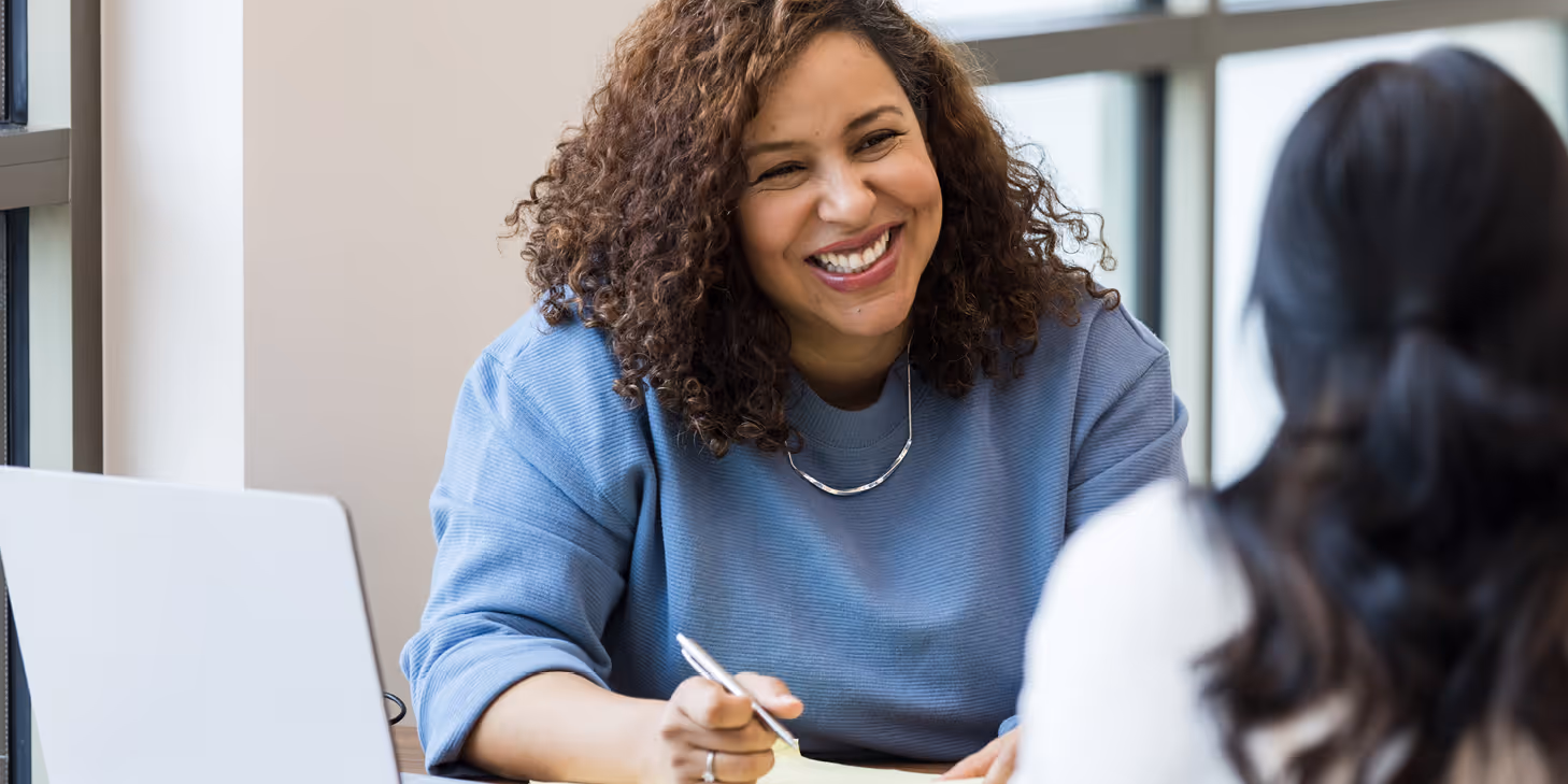 Smiling professional woman in a casual meeting with a colleague, holding a pen and sitting at a desk with a laptop in a bright office setting.