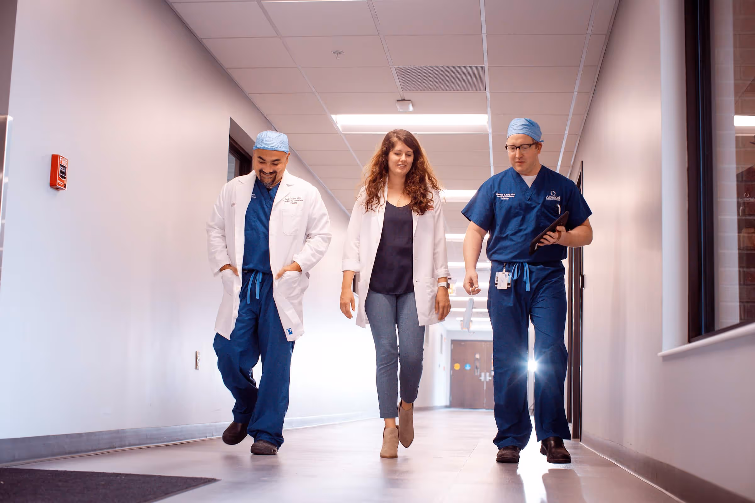 Three medical professionals walking down a hospital hallway, two men in blue scrubs with white coats and a woman in a white coat and casual pants.