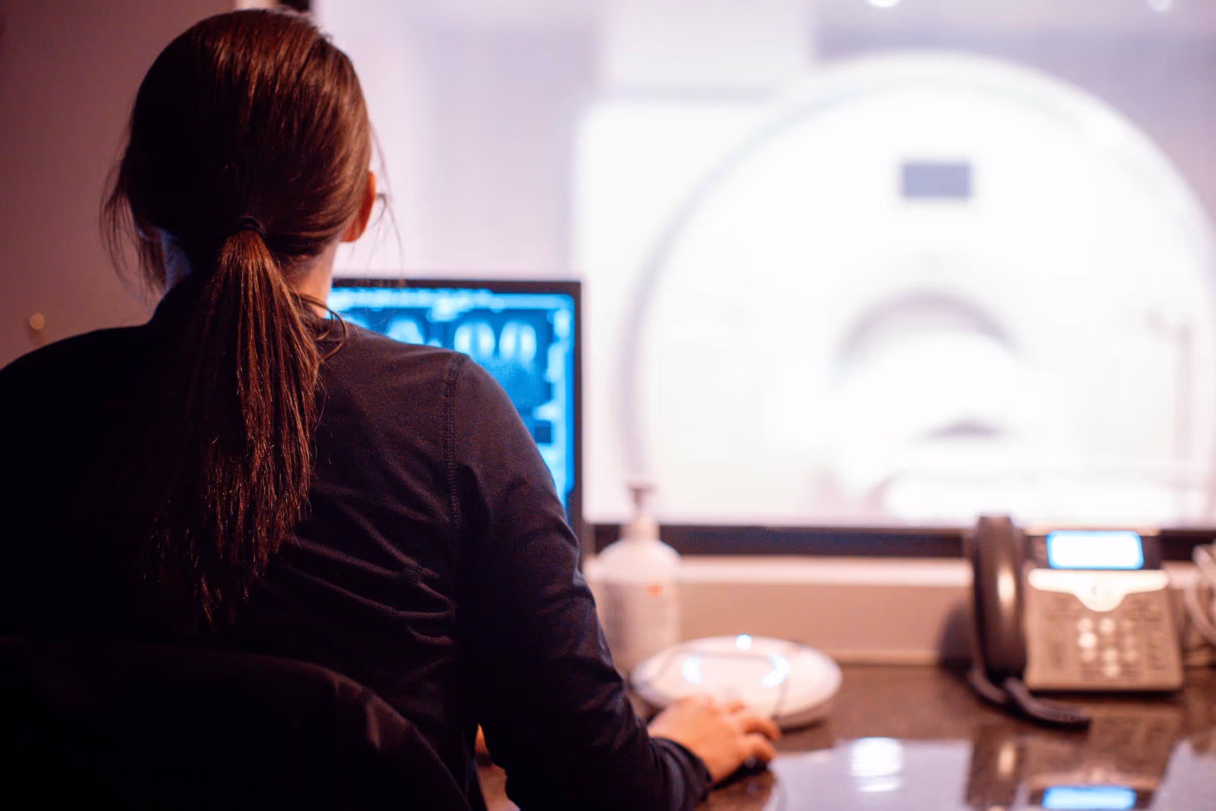 Technician with hair tied back operating computer in front of MRI machine visible through window.