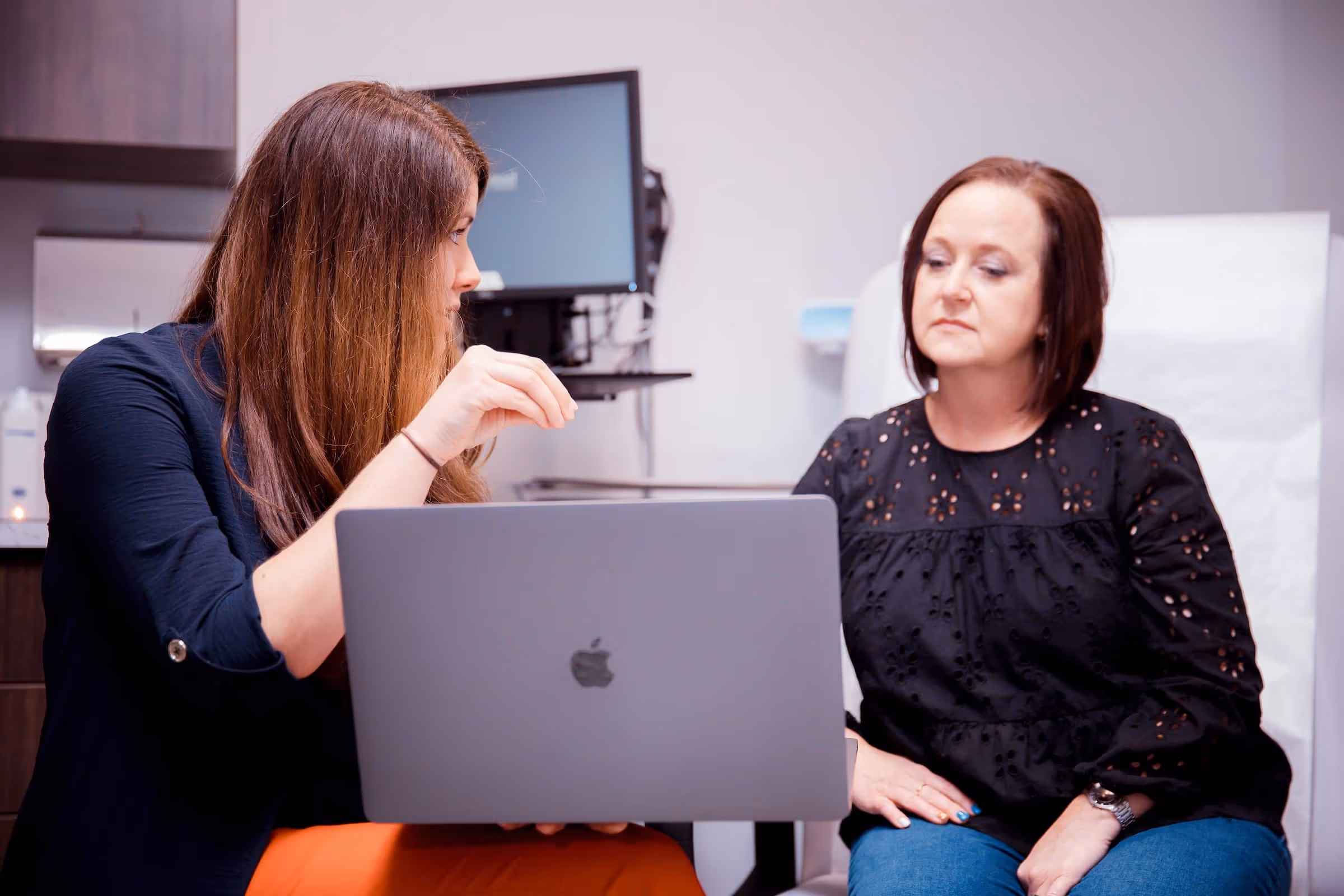 Two women in a consultation setting, one showing something on a laptop to the other in a medical office.