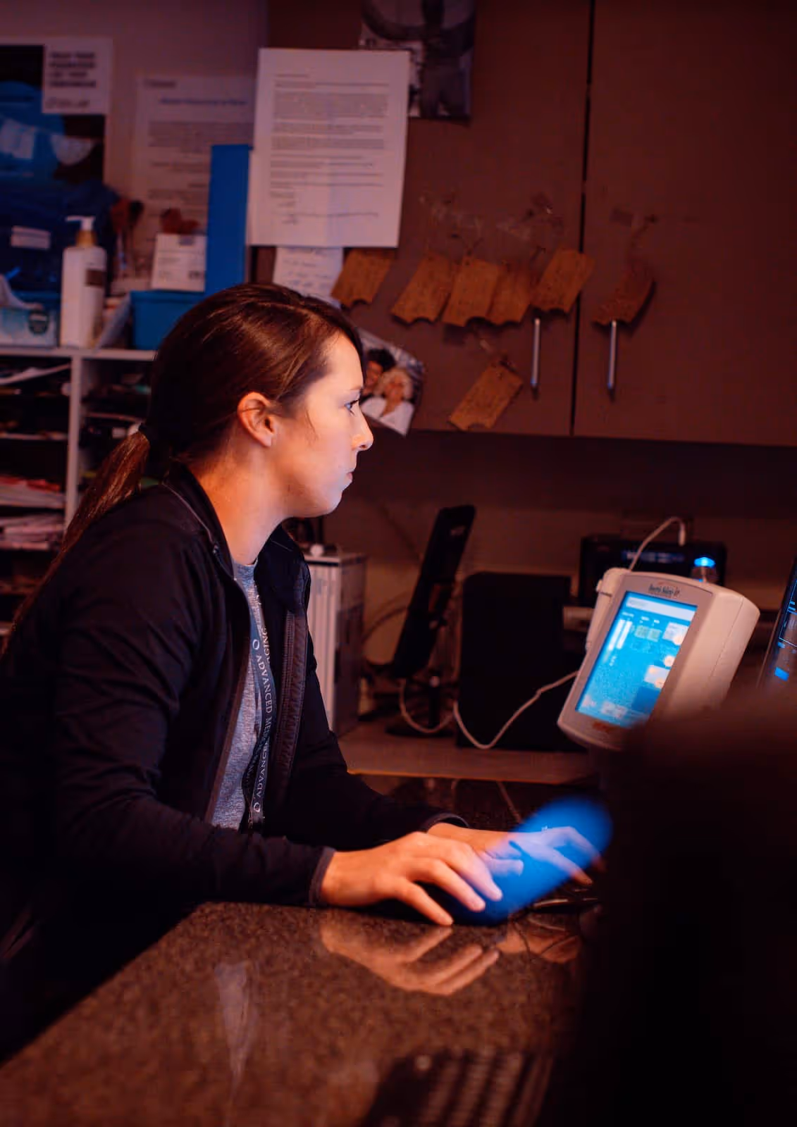 Woman with dark hair in a ponytail using a computer mouse and looking at a monitor in a dimly lit office.