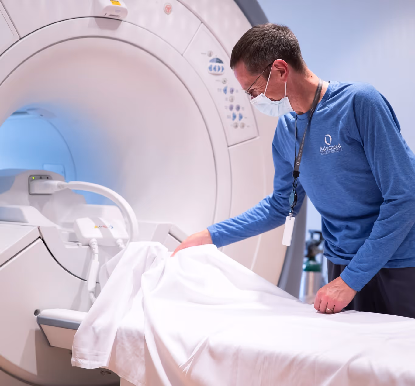 Technician wearing a mask and blue shirt prepares the patient table by covering it with a white sheet in front of an MRI machine.