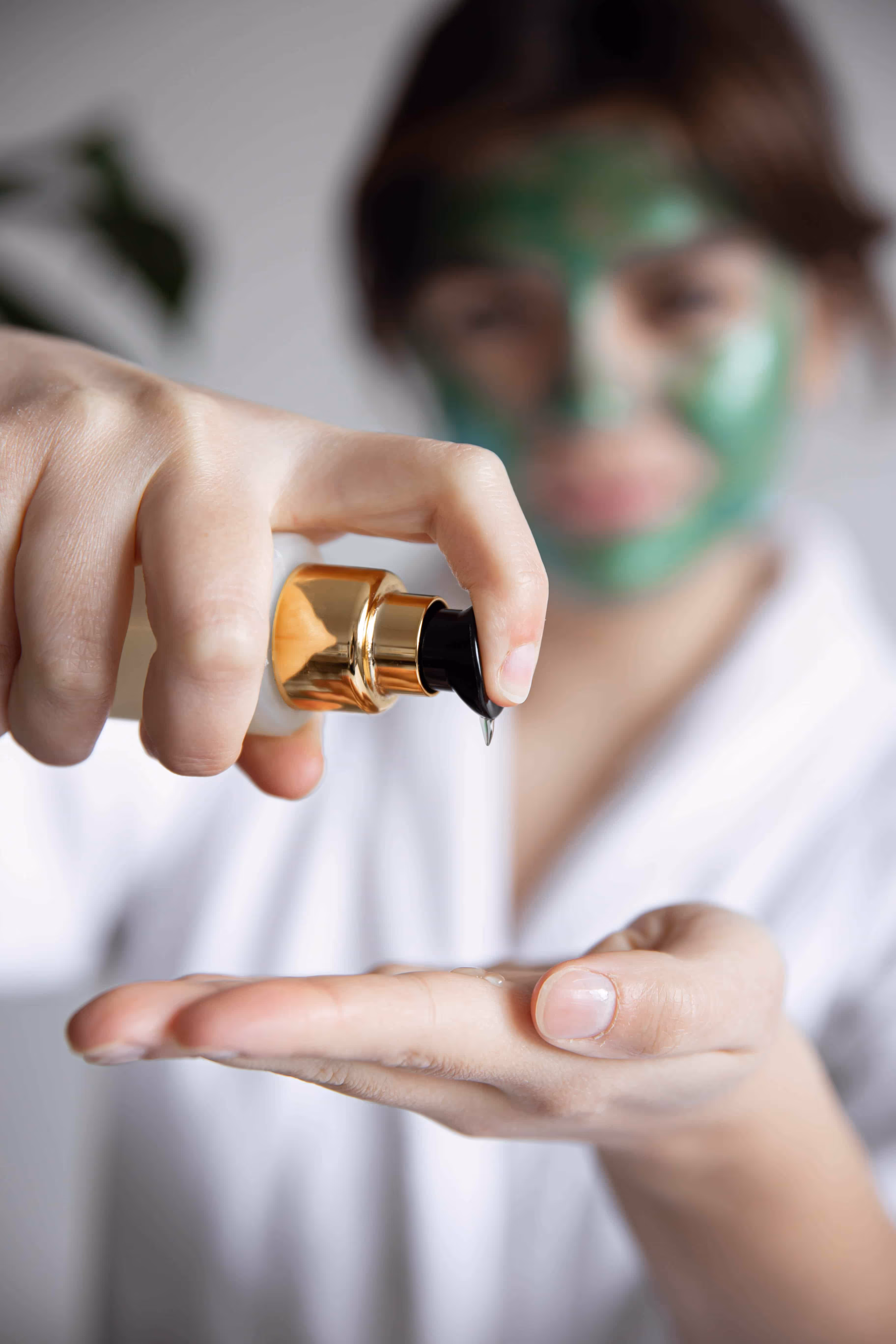 Person with green facial mask dispensing serum or lotion onto their palm from a pump bottle.