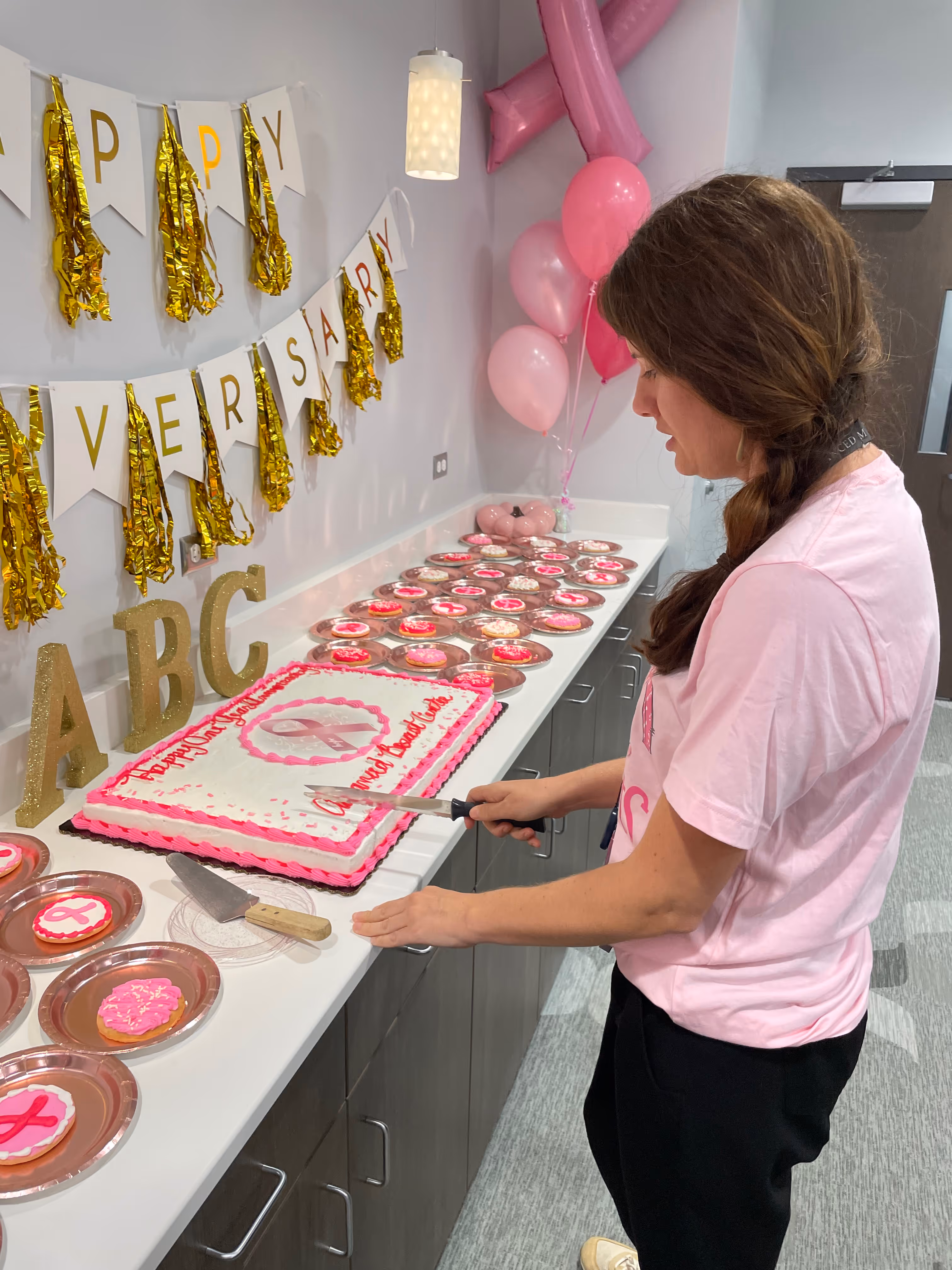 Woman in pink shirt cutting a large anniversary cake decorated with a pink ribbon on a counter with pink-themed cookies and balloons.