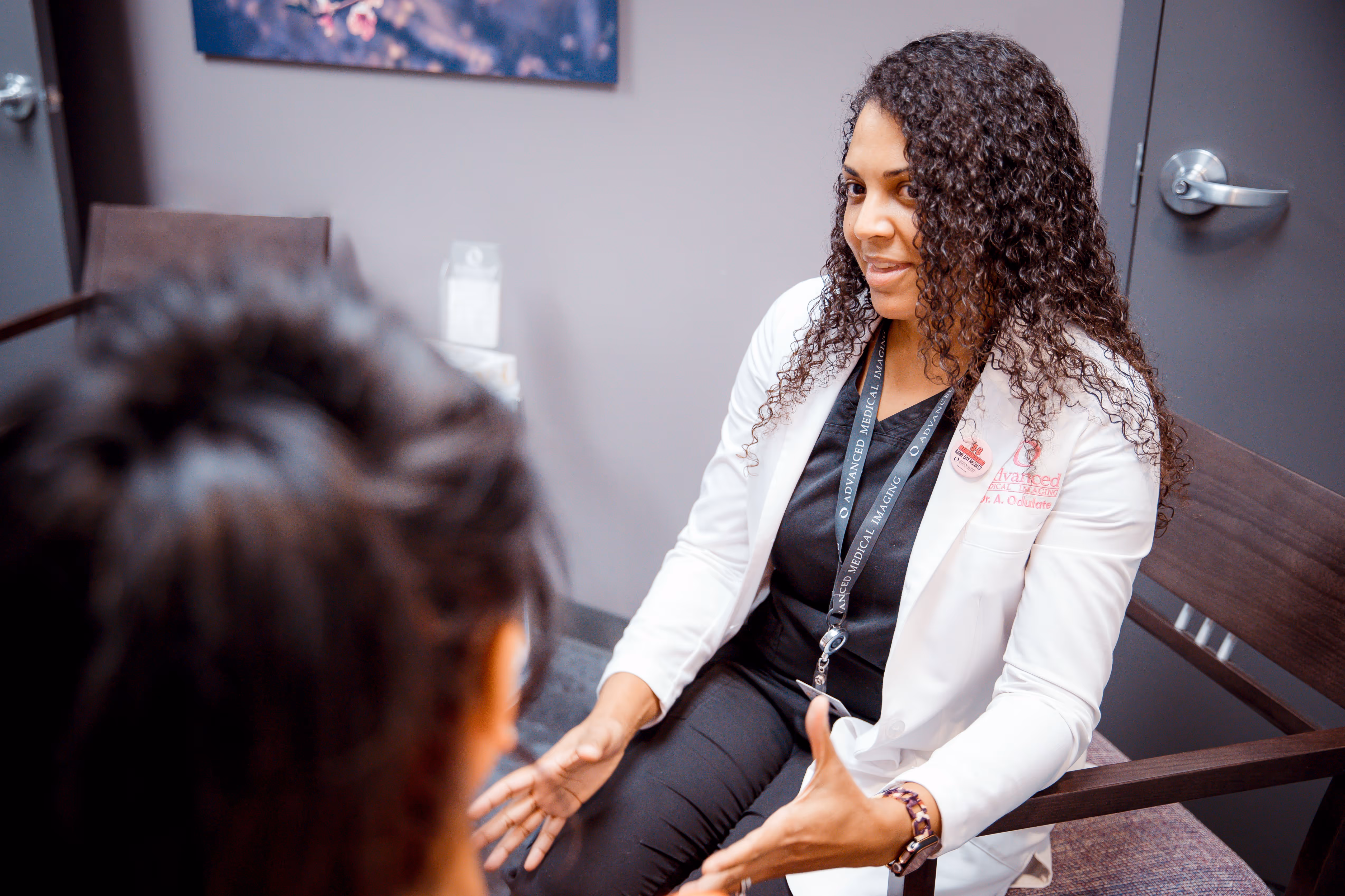 Female doctor with curly hair in a white coat talking to a patient in a medical office.