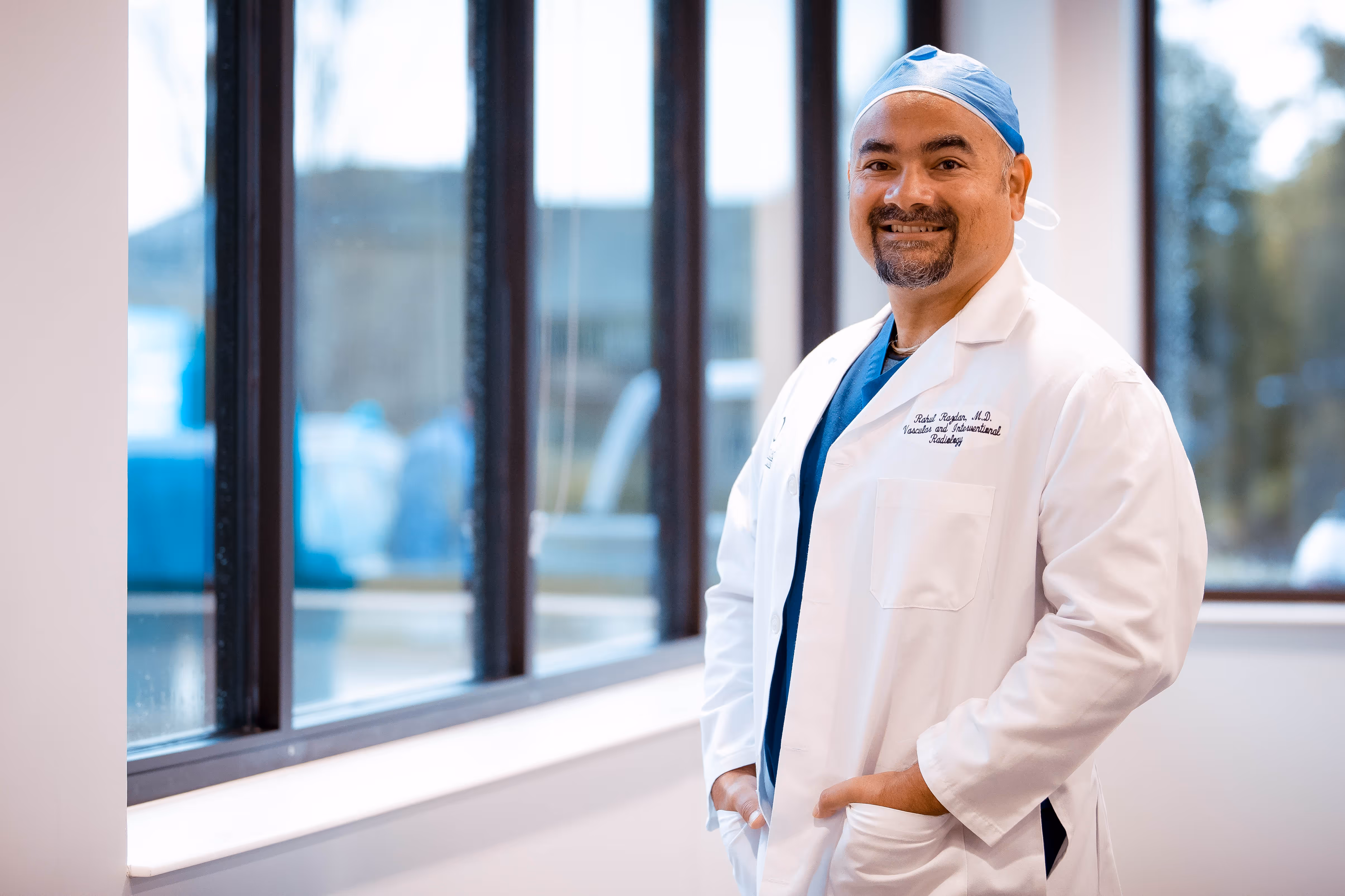 Smiling male doctor wearing a white lab coat and blue scrub cap standing indoors by large windows.