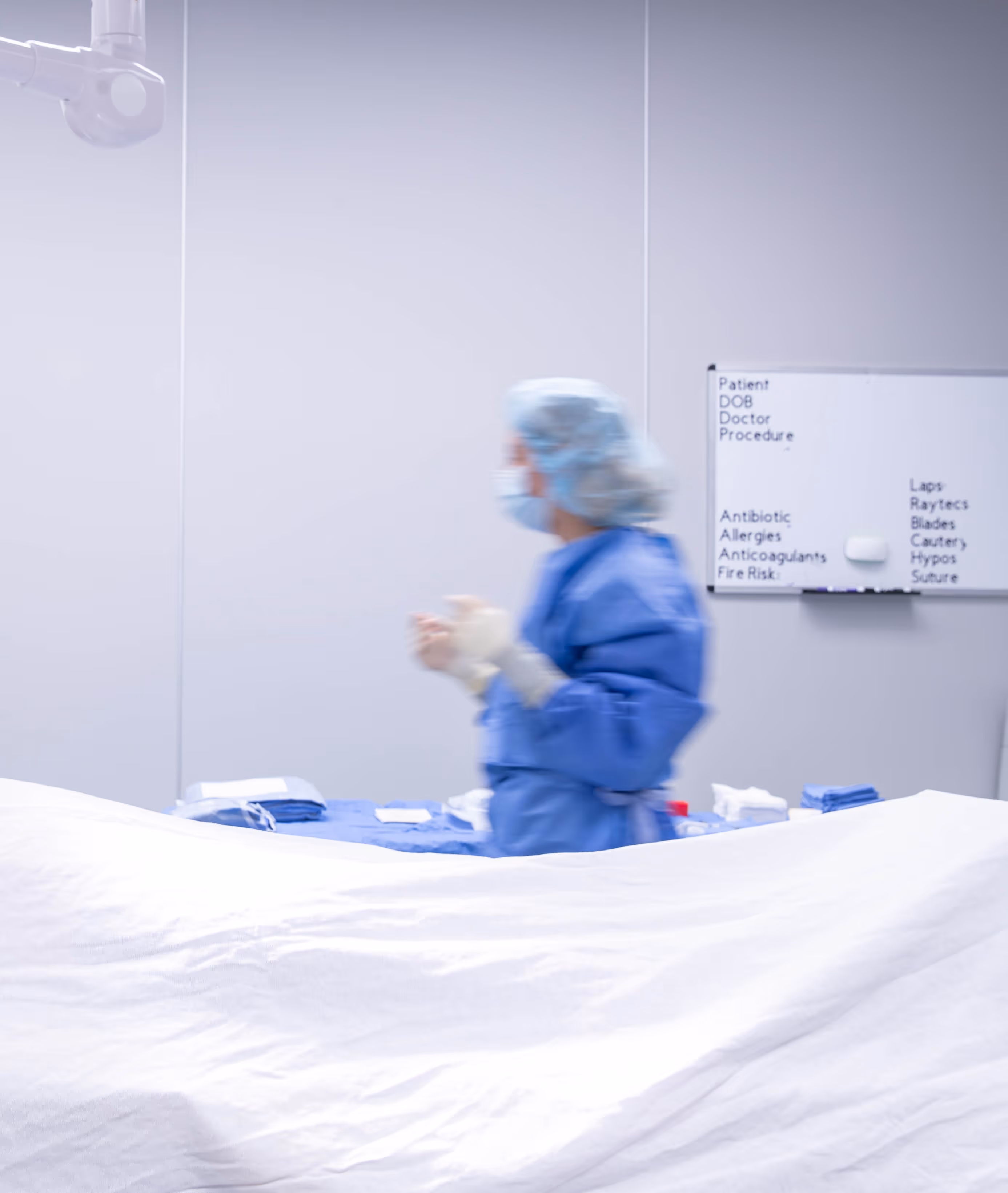 Medical professional in blue scrubs and hairnet standing in a clinical setting with a blurred whiteboard listing medical terms in the background.