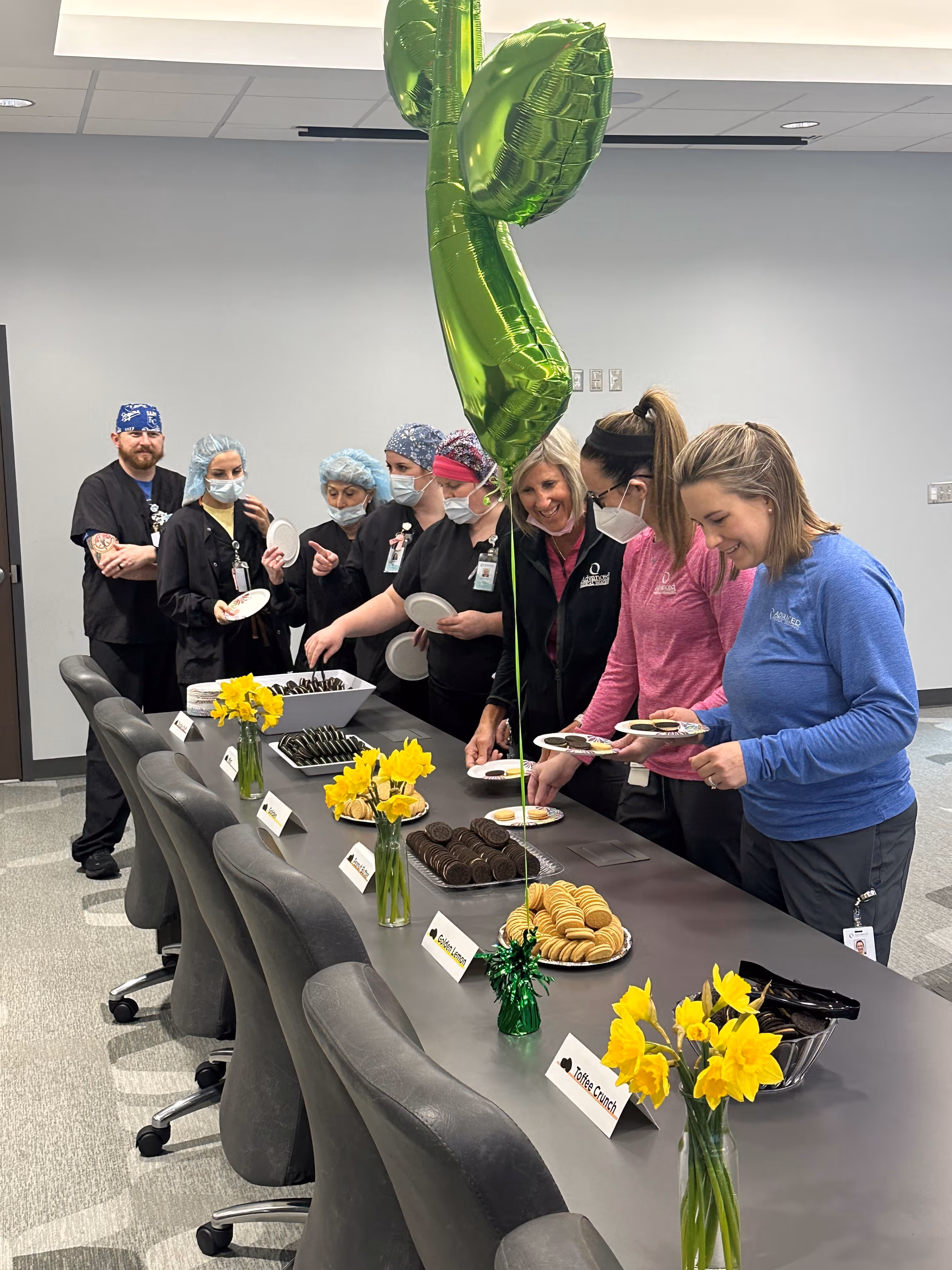 Group of healthcare workers in scrubs and masks gathered around a table with plates and various types of cookies labeled with flavor names, decorated with yellow daffodils and green balloons.