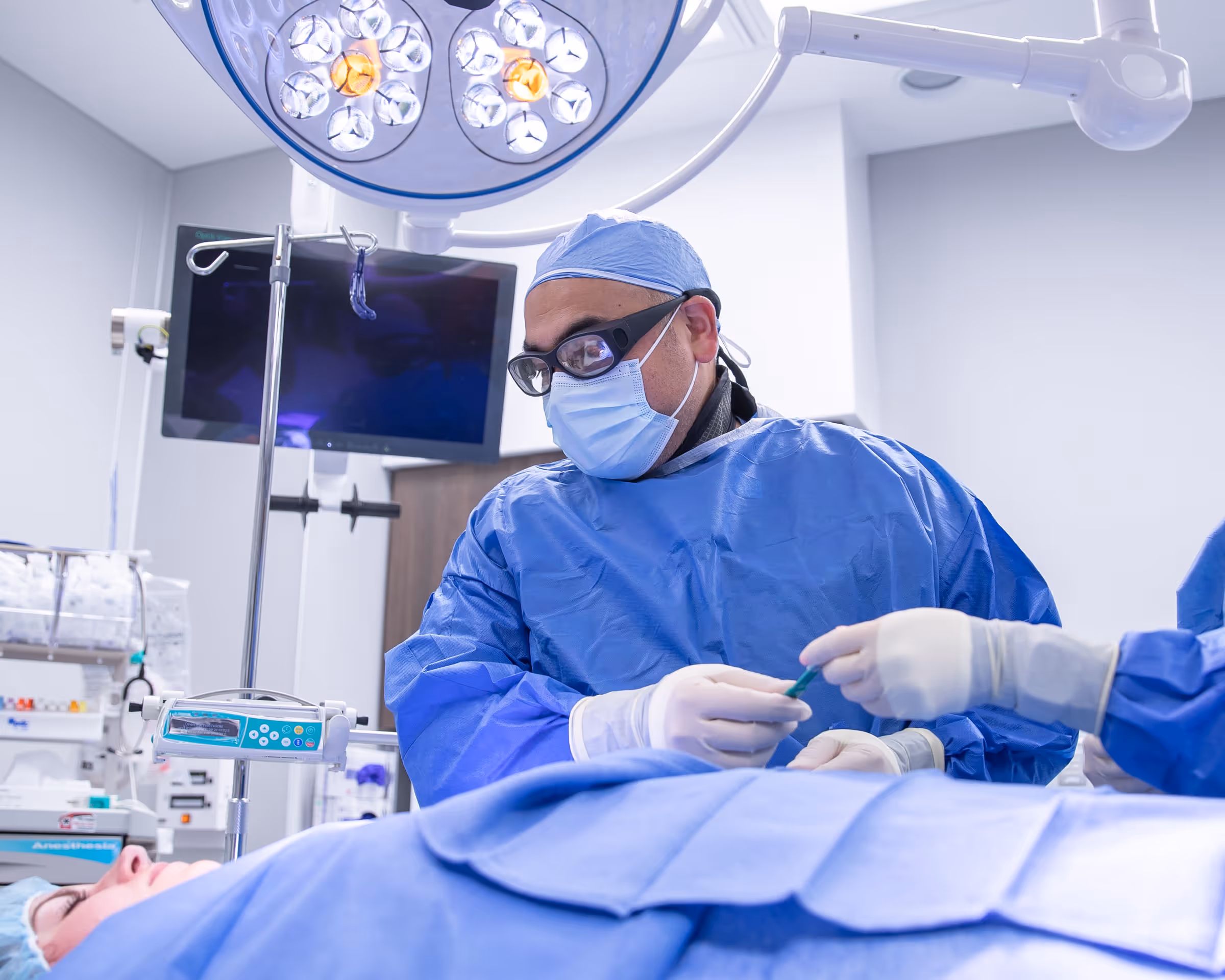 Surgeon in blue scrubs, mask, and protective glasses preparing for surgery in an operating room with patient covered in blue drapes.